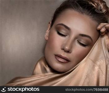 Closeup Portrait of a Beautiful Woman with Evening Makeup Wrapped in a Golden Silk Shawl over Beige Background. Luxury Fashion Look.