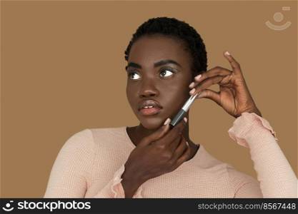 Closeup portrait of a beautiful young black woman with short Afro hair, light makeup and lipstick posing by herself holding a lipstick in both of her hands inside a studio with a pecan background.