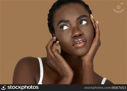 Closeup portrait of a cute young black woman with short Afro hair, light makeup and lipstick posing by herself inside a studio with a pecan background resting the palms of her hands on her face.