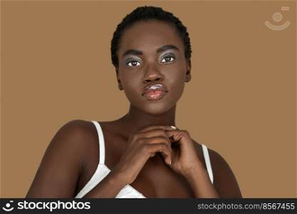 Closeup portrait of a cute young black woman with short Afro hair, light makeup and lipstick posing by herself inside a studio with a pecan background wearing a white strapped dress.