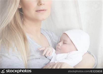 Closeup portrait of a mother standing near the window with cute sleeping baby in hands, happy family, new life concept