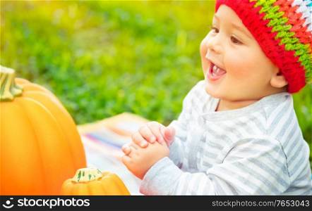 Closeup portrait of a nice cheerful baby boy laughing and enjoying time spending outdoors, sitting near two pumpkins, traditional symbol of Thanksgiving day