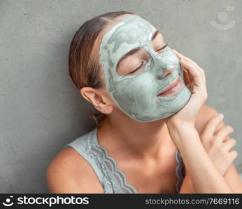 Closeup portrait of a nice female with a mask of blue clay on her face over gray background, enjoying day spa, closing eyes of pleasure, beauty and skin care