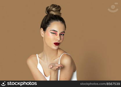 Closeup portrait of a playful young Latina with long dark blond hair, eye makeup and lipstick sitting by herself on top of a table inside a studio with a pecan background wearing a white strap top.