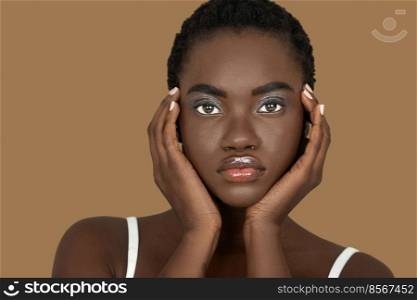 Closeup portrait of a sensual young black woman with short Afro hair, light makeup and lipstick posing by herself inside a studio with a pecan background resting the palms of her hands on her face.