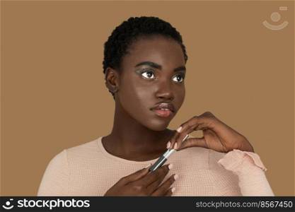 Closeup portrait of a serene young black woman with short Afro hair, light makeup and lipstick posing by herself holding a lipstick in both of her hands inside a studio with a pecan background.