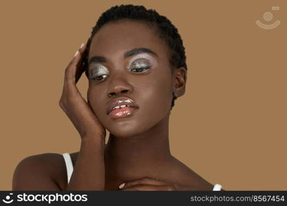 Closeup portrait of a serene young black woman with short Afro hair, light makeup and lipstick posing by herself inside a studio with a pecan background resting her palm of her hand on her face.