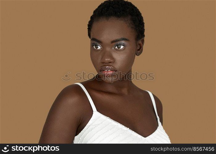 Closeup portrait of a serene young black woman with short Afro hair, light makeup and lipstick posing by herself inside a studio with a pecan background wearing a white strapped dress.