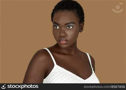 Closeup portrait of a serene young black woman with short Afro hair, light makeup and lipstick posing by herself inside a studio with a pecan background wearing a white strapped dress.