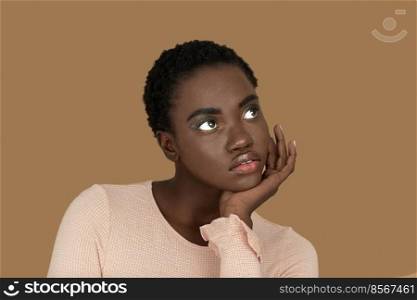 Closeup portrait of a serene young black woman with short Afro hair, light makeup and lipstick posing by herself resting her head on the palm of her hand inside a studio with a pecan background.