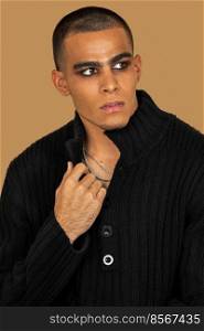 Closeup portrait of a serene young Latin man with short hair, light makeup and eyeshadow sitting by himself inside a studio with a pecan background wearing black jeans and a black sweater.