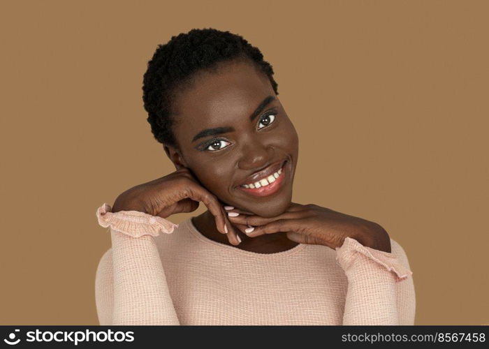 Closeup portrait of a smiling young black woman with short Afro hair, light makeup and lipstick posing by herself resting her head on the palm of her hand inside a studio with a pecan background.