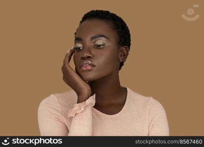 Closeup portrait of an attractive young black woman with short Afro hair, light makeup and lipstick posing by herself resting her head on the palm of her hand inside a studio with a pecan background.