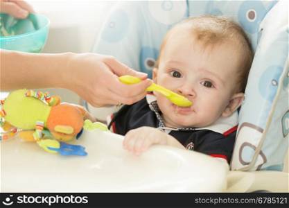 Closeup portrait of baby boy sitting behind table at highchair and eating from spoon