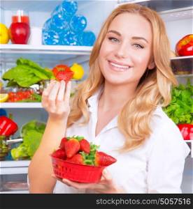 Closeup portrait of beautiful female eating strawberry, open fridge full of fruits and vegetables, dieting and healthy eating concept