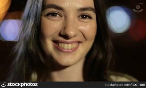 Closeup portrait of beautiful young brunette woman smiling at night over streetlights bokeh background. Joyful young female posing and looking at camera with perfect toothy smile in blurry dimmed lights of night city.