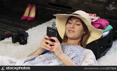 Closeup portrait of beautiful young woman in panama hat lying on the floor and sending a text message on smartphone at home. Attractive female holding mobile phone while lying on back, with head on packed travel case, getting ready for new journey.