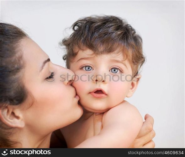 Closeup portrait of cheerful young mother kissing in cheek her precious little son, isolated on gray background, happy family life