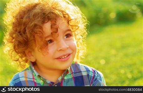 Closeup portrait of cute baby boy with curly hair on green field, having fun outdoors, enjoying summer vacation, happy childhood concept