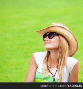 Closeup portrait of cute blond female wearing sunglasses and straw hat sitting down on green grass, having fun outdoors, summer vacation concept
