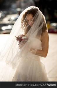 Closeup portrait of cute curly bride with long veil looking at bouquet
