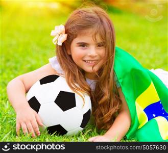 Closeup portrait of cute little football fan lying down on fresh green grass with ball and big Brazil flag, happy supporter of Brazilian football team