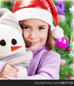 Closeup portrait of cute little girl holding snowman soft toy over Christmas tree background, wearing red Santa hat&#xA;