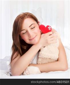 Closeup portrait of cute young girl lying down in her bed at home, closing eyes of pleasure, receive soft bear toy with red heart as gift for Valentine day