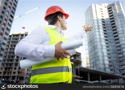 Closeup portrait of foreman with blueprints pointing hand at high building