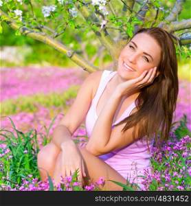 Closeup portrait of nice dreamy girl sitting down on pink flowers field in apple tree garden, watching for first spring blossom, beauty of nature concept