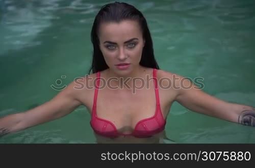 Closeup portrait of sexy young woman wearing pink bikini with wet hair looking at camera while walking out of outdoor swimming pool at tropical resort - video in slow motion