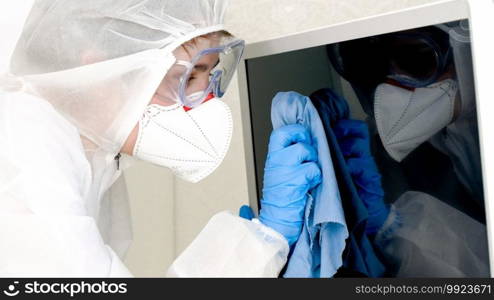 Closeup portrait of woman in medical suit and protective mask doing housework and desinfecting television and furniture at home. Phobia of covid-19 and coronavirus panic.. Closeup portrait of woman in medical suit and protective mask doing housework and desinfecting television and furniture at home. Phobia of covid-19 and coronavirus panic