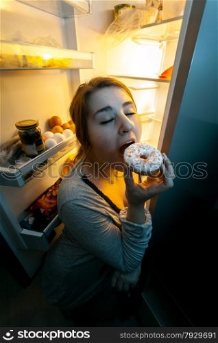 Closeup portrait of woman in pajamas eating donut on kitchen at night