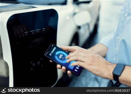 Closeup progressive businessman look at EV car&rsquo;s battery status application on smart phone screen at public parking charging station with power cable plug and renewable energy-powered electric vehicle. Closeup progressive idea hand holding smart phone display EV&rsquo;s batter status.
