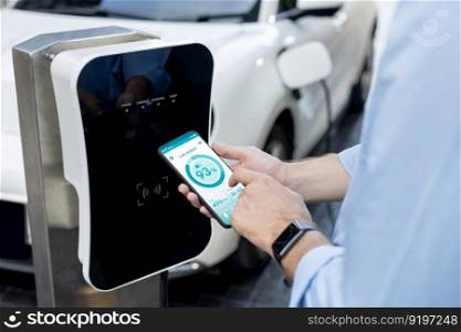 Closeup progressive businessman look at EV car&rsquo;s battery status application on smart phone screen at public parking charging station with power cable plug and renewable energy-powered electric vehicle. Closeup progressive idea hand holding smart phone display EV&rsquo;s batter status.