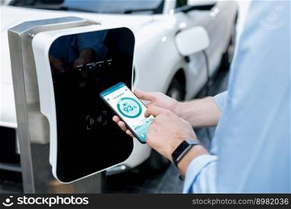 Closeup progressive businessman look at EV car’s battery status application on smart phone screen at public parking charging station with power cable plug and renewable energy-powered electric vehicle. Closeup progressive idea hand holding smart phone display EV’s batter status.