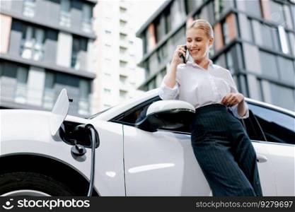 Closeup progressive businesswoman talking on smartphone, leaning electric car at charging station with residential condo apartment in background. Eco friendly rechargeable EV car concept.. Closeup progressive woman talking on the phone, leaning on electric vehicle.