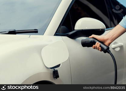 Closeup progressive man holding EV charger plug from public charging station for electric vehicle with background of residential building as concept eco-friendly sustainability energy car concept.. Closeup progressive businessman plugs charger plug from charging station to EV.