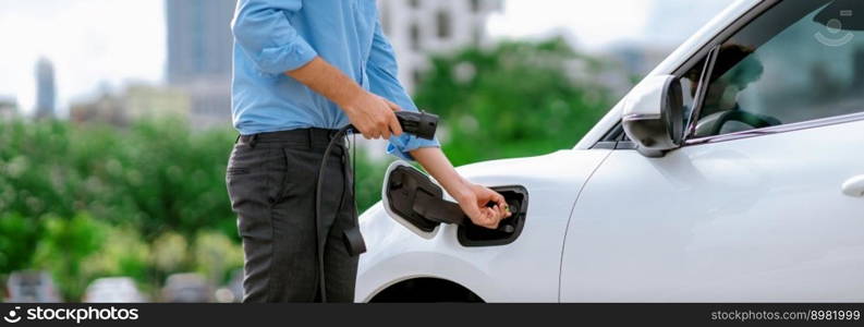 Closeup progressive man holding EV charger plug from public charging station for electric vehicle with background of residential building as concept eco-friendly sustainability energy car concept.. Closeup progressive businessman plugs charger plug from charging station to EV.