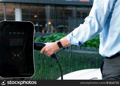 Closeup progressive man holding EV charger plug from public charging station for electric vehicle with background of residential building as concept eco-friendly sustainability energy car concept.. Closeup progressive businessman plugs charger plug from charging station to EV.