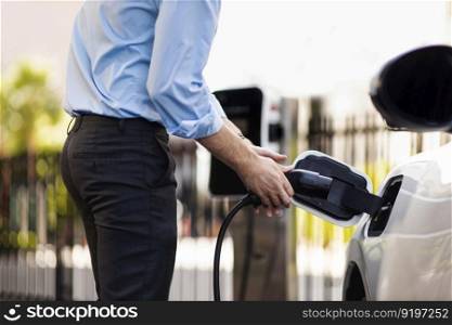 Closeup progressive man holding EV charger plug from public charging station for electric vehicle with background of residential building as concept eco-friendly sustainability energy car concept.. Closeup progressive businessman plugs charger plug from charging station to EV.