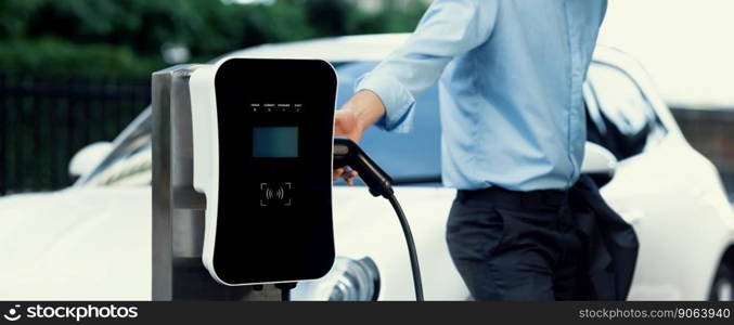 Closeup progressive suit-clad businessman with his electric vehicle recharge his car on public charging station in modern city with power cable plug and renewable energy-powered electric vehicle.. Closeup progressive businessman with electric vehicle at charging station.