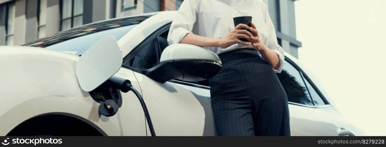 Closeup progressive suit-clad businesswoman with her electric vehicle recharge her car on public charging station in modern city with power cable plug and renewable energy-powered electric vehicle.. Closeup progressive businesswoman with electric vehicle at charging station.