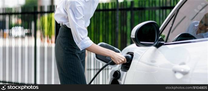 Closeup progressive suit-clad businesswoman with her electric vehicle recharge her car on public charging station in modern city with power cable plug and renewable energy-powered electric vehicle.. Closeup progressive businesswoman with electric vehicle at charging station.
