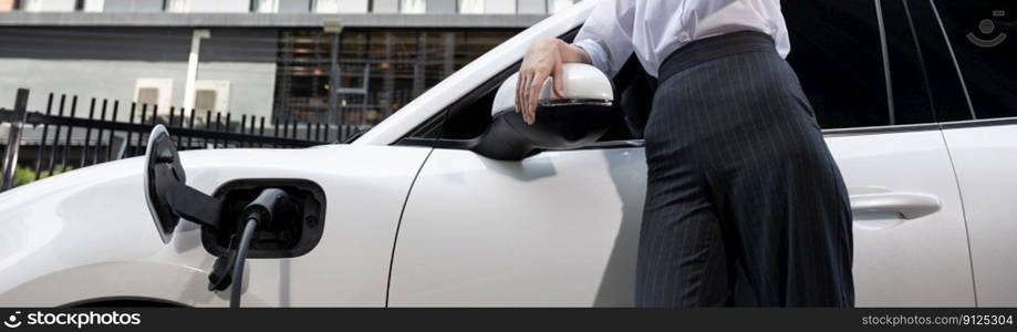 Closeup progressive suit-clad businesswoman with her electric vehicle recharge her car on public charging station in modern city with power cable plug and renewable energy-powered electric vehicle.. Closeup progressive businesswoman with electric vehicle at charging station.