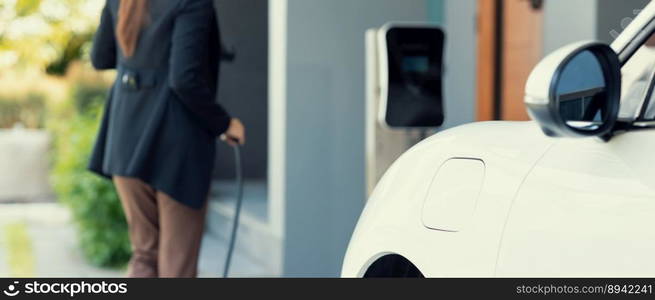 Closeup progressive woman install cable plug to her electric car with home charging station. Concept of the use of electric vehicles in a progressive lifestyle contributes to clean environment.. Closeup progressive woman recharge her EV car at home charging station.