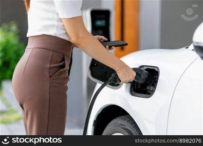 Closeup progressive woman install cable plug to her electric car with home charging station. Concept of the use of electric vehicles in a progressive lifestyle contributes to clean environment.. Closeup progressive woman recharge her EV car at home charging station.