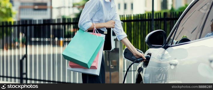 Closeup recharging EV car at charging station with businesswoman carrying shopping bag. Progressive lifestyle of a man in city with ecological concern for clean electric energy driven car ideal.. Closeup EV car with businesswoman carrying shopping bag at charging station.