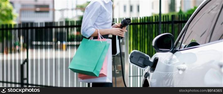 Closeup recharging EV car at charging station with businesswoman carrying shopping bag. Progressive lifestyle of a man in city with ecological concern for clean electric energy driven car ideal.. Closeup EV car with businesswoman carrying shopping bag at charging station.