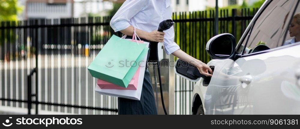 Closeup recharging EV car at charging station with businesswoman carrying shopping bag. Progressive lifestyle of a man in city with ecological concern for clean electric energy driven car ideal.. Closeup EV car with businesswoman carrying shopping bag at charging station.